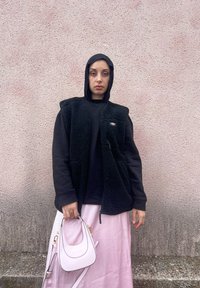 Μαύρο γιλέκο από φλις πάνω από μαύρο τοπ και μακριά ροζ σατέν φούστα, κρατώντας μια μικρή ροζ τσάντα με χρυσές λεπτομέρειες. Το φόντο είναι μια υφασμάτινη ροζ τοίχος.