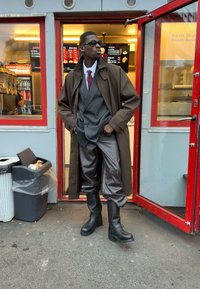 Una persona vestita con un trench marrone sopra un completo grigio, pantaloni grigi a fantasia e stivali neri con tacco si trova all'esterno di un negozio di kebab con finestre rosse.