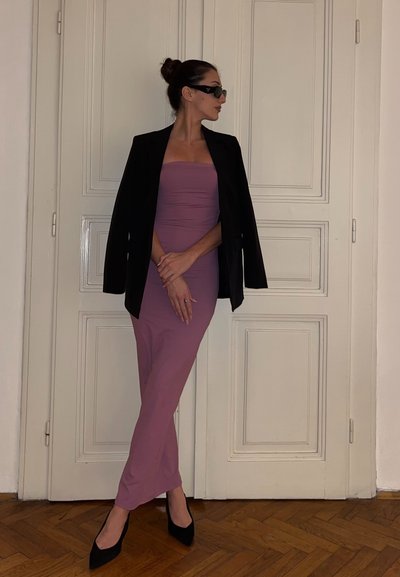 Vestido ajustado sin tirantes en color malva, con una chaqueta negra encima, combinado con tacones negros de punta; unas gafas de sol completan el conjunto contra una pared blanca.