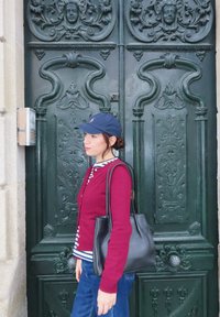 Uma mulher vestindo um cardigã bordô com punhos listrados, jeans azuis e uma bolsa preta de ombro está em pé ao lado de uma porta verde ornamentada com esculturas detalhadas.