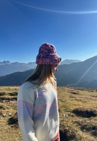 Pleteni vijolični in roza klobuk z zavihanjem, nošen z belim puloverjem, ki ima pastelne geometrijske vzorce in dolge lase, ki so vidne.