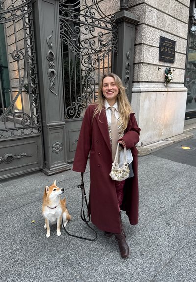 Abrigo burdeos, corbata crema sobre camisa a cuadros, bolso estampado, botas oscuras; un Shiba Inu está sentado al lado, sobre una acera de piedra con una verja ornamentada de fondo.