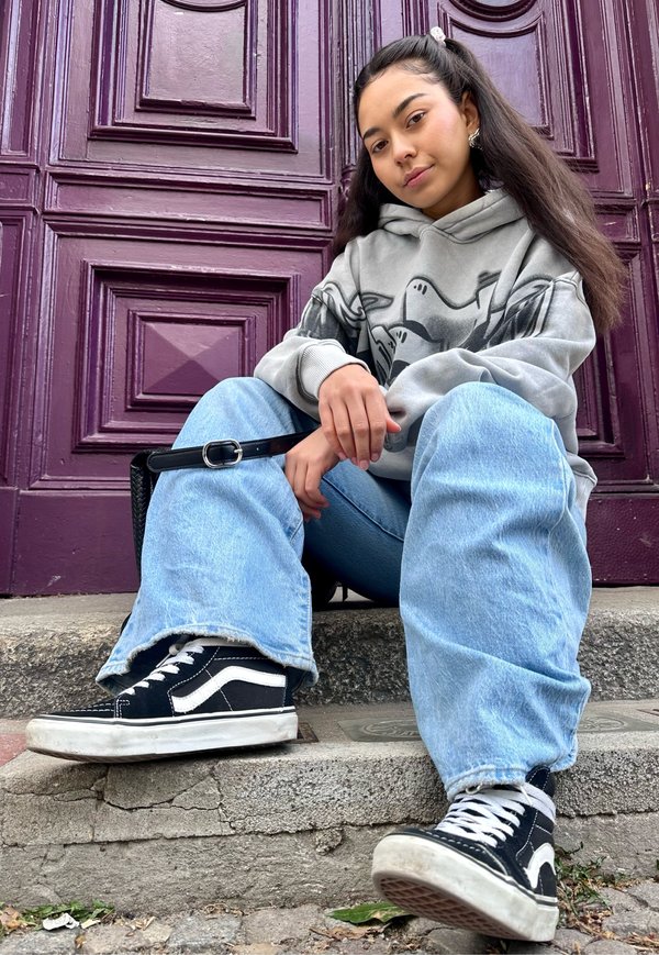 Joven con cabello largo, con sudadera gris con capucha y vaqueros anchos, sentada en escalones de piedra frente a una puerta púrpura con paneles.