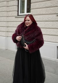 Γυναίκα με κόκκινα μαλλιά που φοράει ένα βουργουνδί τζάκετ από ψεύτικη γούνα και μαύρη φούστα, κρατώντας μια μαύρη καπιτονέ τσάντα με αλυσίδα.