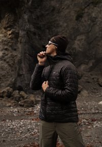 Černá zateplená bunda s prošitým vzorem, na zip vpředu a nastavitelnou kapucí. Nošena s hnědou čepicí a šedými kalhotami venku.