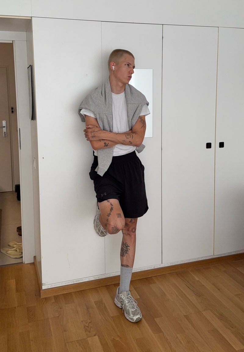 Jeune personne avec des tatouages, portant un t-shirt blanc, un short noir, des baskets grises, debout à l'intérieur contre une armoire blanche, les bras croisés.