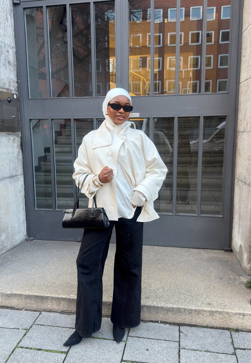 Persona, valkājot baltu jaku, galvas lakatu un melnas saulesbrilles, turot melnu somu, stāvot pie pelēkām metāla durvīm ar stikla paneļiem.