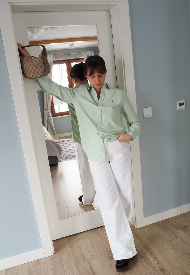 Femme en chemise vert clair et pantalon blanc appuyée contre le cadre d'une porte, tenant un sac à main beige, debout sur un sol en bois avec un miroir derrière elle.
