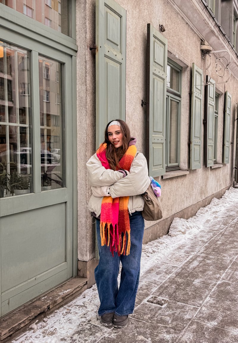 Jeune femme portant une écharpe colorée, une veste beige et un jean, debout sur un trottoir enneigé à côté d'un bâtiment avec des volets et des fenêtres verts.