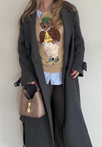 Manteau gris porté sur un pull beige en tricot avec un motif d'ours, associé à une chemise bleu clair, un pantalon foncé et un sac à main beige avec des accents dorés.