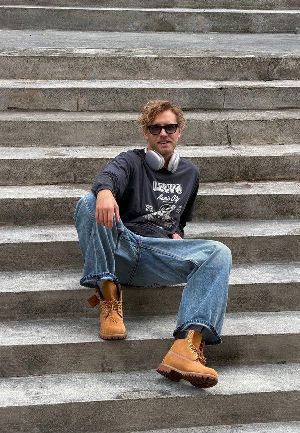 Hombre con gafas de sol, auriculares, camisa de manga larga oscura, vaqueros y botas beige sentado en una escalera de hormigón al aire libre.