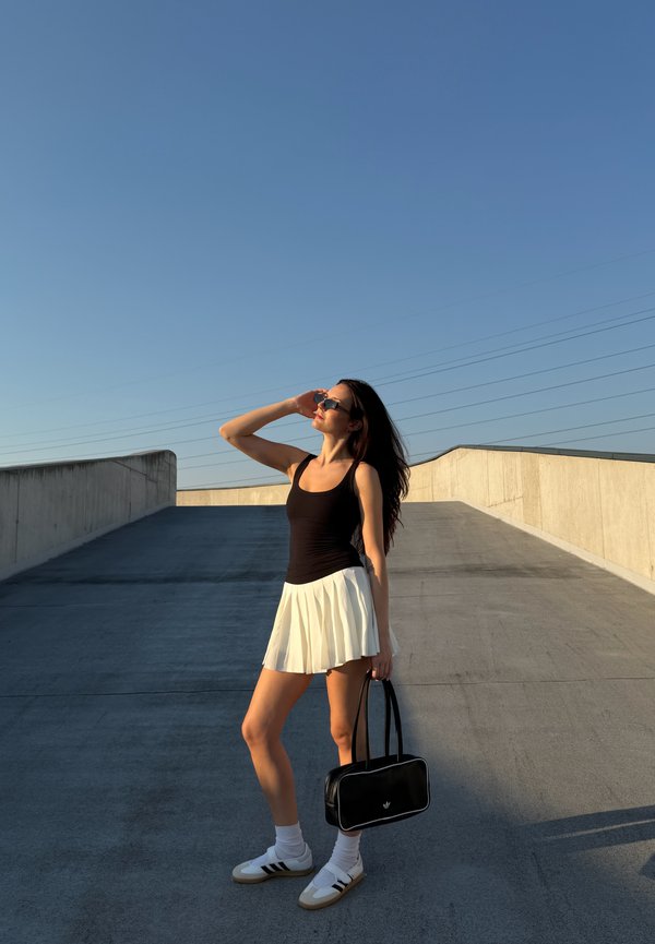 Femme en débardeur noir et jupe blanche tenant un sac noir, debout sur une pente pavée sous un ciel bleu clair, protégeant ses yeux avec des lunettes de soleil.
