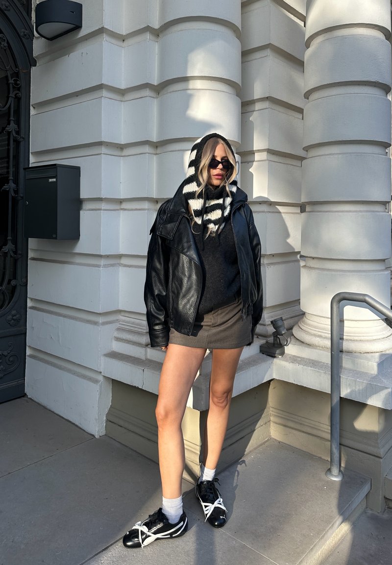 Schwarze Lederjacke, gestrickter schwarz-weißer gestreifter Schal, schwarzer Pullover, grauer Rock, schwarze Schuhe mit weißen Schnürsenkeln, stehend vor einer weißen Wand.