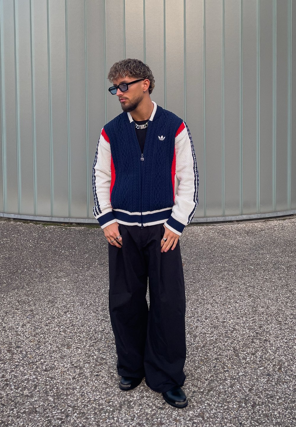 Cardigan tricotat cu fermoar, cu blocuri de culoare roșu, alb și bleumarin; textură groasă; pantaloni negri cu picioare evazate; ochelari de soare rotunzi; inele mari.