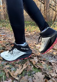 Scarpe da trail GORE-TEX con tomaia bianca e nera, superficie testurizzata, tallone rinforzato e suola rosa, indossate su un pavimento forestale coperto di foglie.