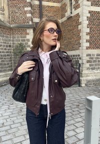 Vinröd skinnjacka med silverdetaljer, två framfickor, kombinerad med en vit skjorta och ljusrosa slips. Svart flätad handväska, mörka jeans.