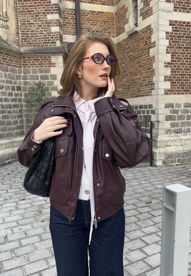 Bordeauxrote Lederjacke mit silbernen Beschlägen, zwei Fronttaschen, kombiniert mit einem weißen Hemd und einer hellrosa Krawatte. Schwarze, gewebte Handtasche, dunkle Jeans.