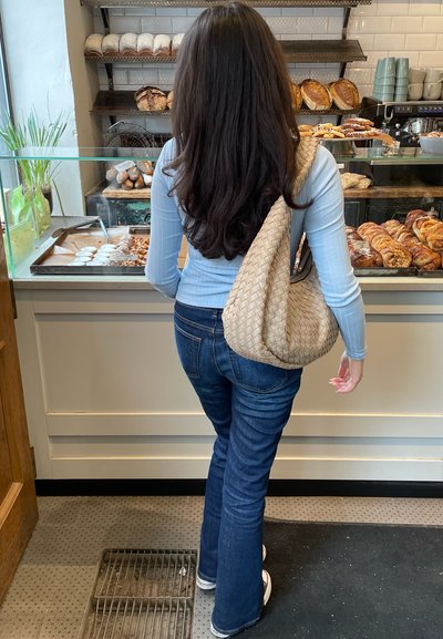 Bolso de hombro de color beige tejido, top de manga larga color azul claro, jeans acampanados de color azul oscuro, de pie frente a una vitrina con una variedad de productos de repostería.