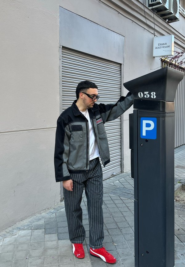 Siv in črn plašč z dvema žepoma, črno progaste hlače in rdeče superge, blizu črnega parkirnega metrskega aparata z modrim oznako.
