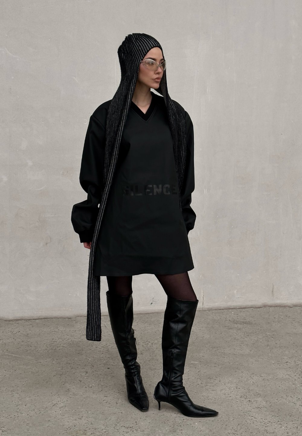 Μαύρο oversized φόρεμα με το κείμενο "SILENCE", μακριά ρίγα μαύρη κουκούλα, διάφανα γυα�λιά, και μαύρες δερμάτινες μπότες μέχρι το γόνατο σε γκρι φόντο.