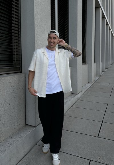Camisa de botones de manga corta de color crema claro sobre una camiseta blanca, combinada con pantalones de pierna ancha negros y zapatillas blancas con acentos beige.