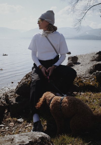 Persona con camiseta blanca de capas, gorro gris y gafas de sol sentada en una roca junto al lago, sosteniendo la correa de un perro marrón rizado que olfatea el suelo.