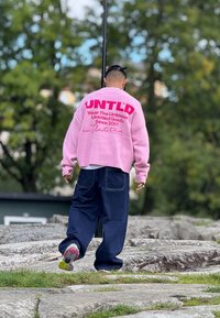 Ružičasti oversized pulover s natpisom "UNTLD", uparen s širokim tamnoplave traperice. Šarene tenisice. Vanjska postavka s zelenilom.