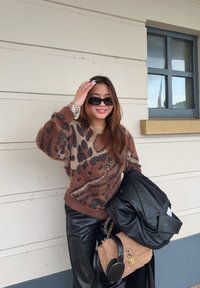 Καφέ και μπεζ πουλόβερ με λεοπάρ σχέδιο, υφασμένο με υφή, σε συνδυασμό με μαύρο δερμάτινο παντελόνι, κρατώντας μια μαύρη δερμάτινη τζάκετ και μια φανερή μπεζ τσάντα.