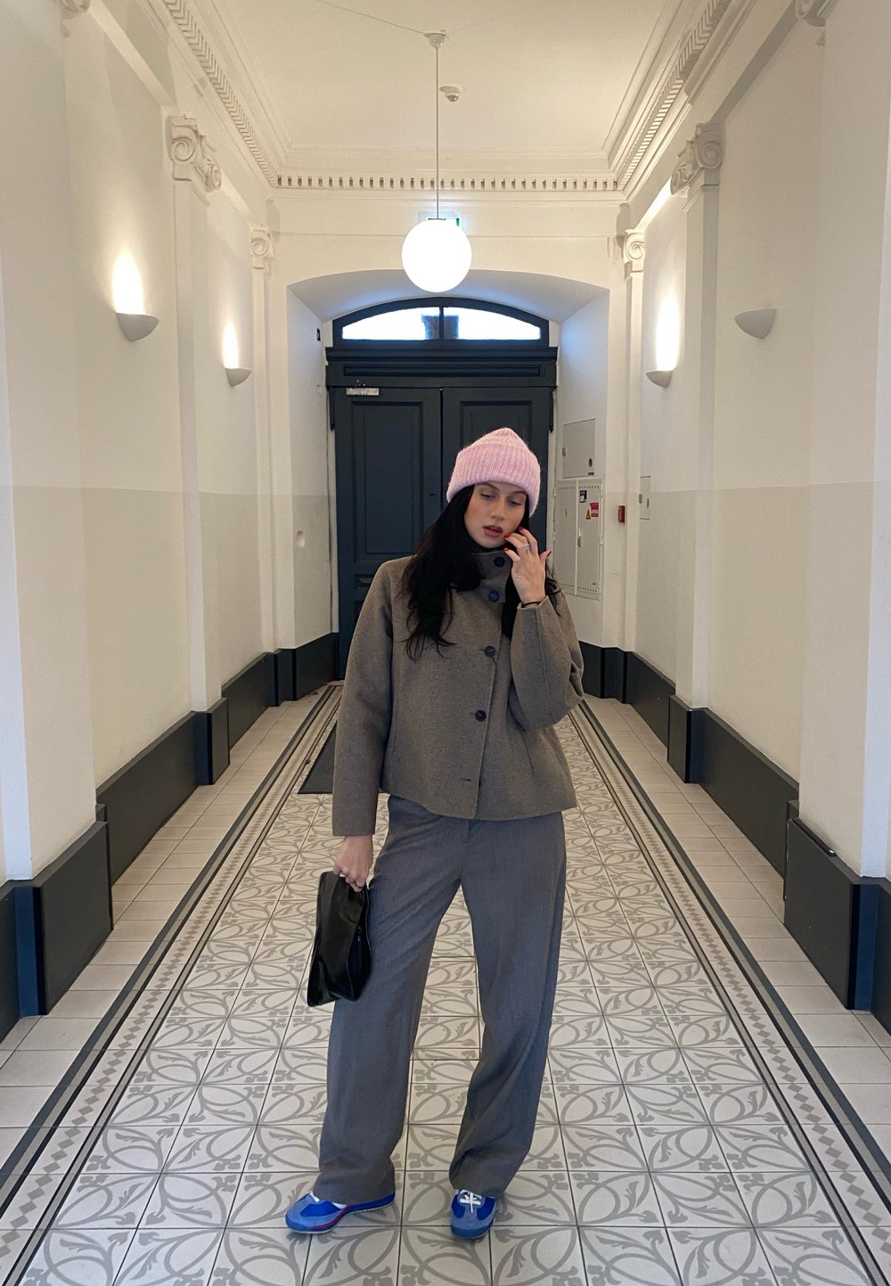 Pelēka šūta jaka ar melnajiem pogām, atbilstošas pelēkas bikses un zilas kurpes. Rozā adīta cepure un melna rokassoma pabeidz apģērbu.