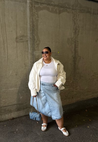 Mujer con chaqueta blanca, camiseta blanca, falda vaquera y zapatos blancos, sosteniendo un pequeño bolso azul, de pie junto a una pared de hormigón.
