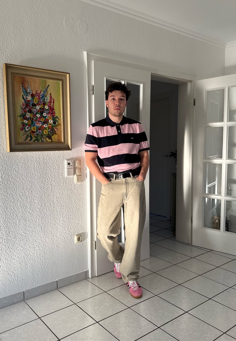 Jeune homme debout dans un cadre de porte, portant un polo rose à rayures noires, un pantalon beige et des baskets roses, sur un sol et un mur carrelés blancs.