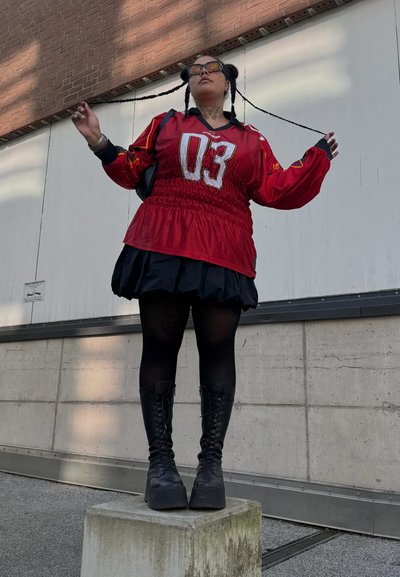 Jersey rojo oversize con impresión "03" sobre una falda negra en capas, medias negras y botas de plataforma con cordones, de pie sobre un bloque de hormigón.
