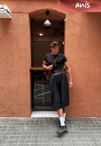 Camiseta negra ajustada, pantalones cortos negros holgados, calcetines blancos y zapatos negros. Cinturón marrón y bolso de hombro. El fondo presenta una pared naranja.