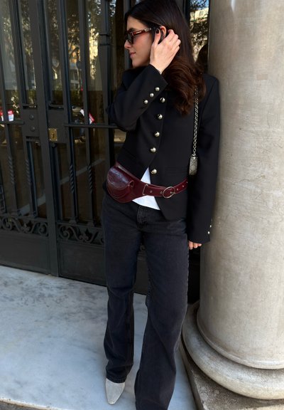 Mujer con blazer negro con botones decorativos, jeans oscuros, riñonera burdeos, zapatos blancos puntiagudos y gafas de sol, de pie junto a una columna.