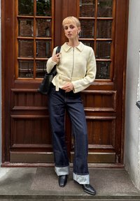 Chaqueta crema con cremallera frontal y cuello, combinada con vaqueros de mezclilla oscuros de pierna ancha con dobladillos remangados y zapatos de cuero negro. Accesorio de bolso negro.