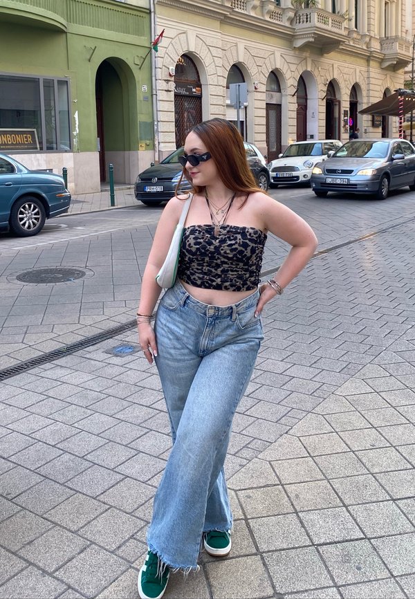 Una persona lleva una camiseta corta de estampado de leopardo negro, jeans de pierna ancha de color azul claro con dobladillos deshilachados y zapatillas verdes, todo contra el fondo de una calle de la ciudad.