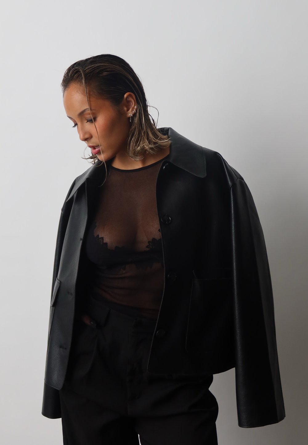 Schwarze Lederjacke mit großem Kragen, getragen über einem transparenten schwarzen Oberteil mit Spitzenakzenten. Das Outfit ist mit schwarzen Hosen kombiniert.