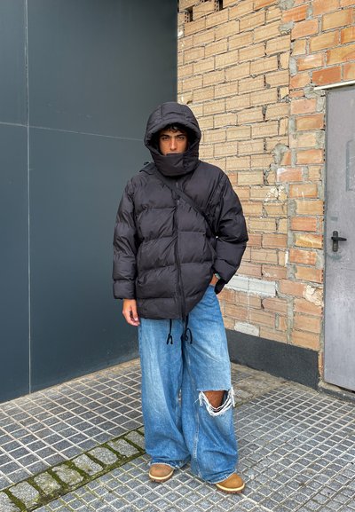 Chaqueta puffer negra con capucha, cierre frontal y dobladillo ajustado, combinada con jeans de mezclilla anchos en un tono azul claro que presentan una rodilla desgastada.