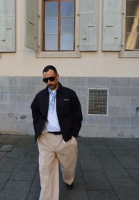 Schwarze Zip-Up-Jacke, weißes T-Shirt, beigefarbene weit geschnittene Hose. Blauer gemusterter Schal um den Hals. Steht auf einem Steinpflaster, mit Fenstern im Hintergrund.