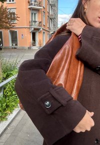 Pruun villane mantel laiade varrukatega ja nööpidega varrukas, hoides läikivat, tekstuurse pruuni nahast kotti. Linna taust värviliste hoonetega.