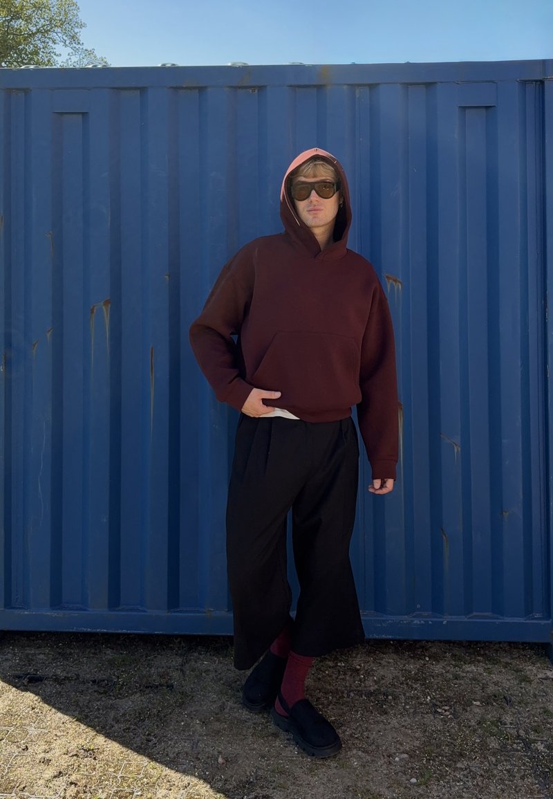 Personne portant un sweat à capuche rouge foncé, un pantalon large noir, des chaussettes bordeaux, des chaussures noires et des lunettes de soleil se tient contre un mur en métal bleu d'un conteneur.