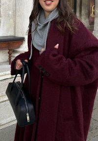 Vinrød, lodden kåpe med store ermer, tre svarte knapper, lag på lag med grå hettegenser, holder en svart skinnhåndveske med glidelås.