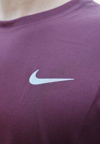 Chemise de sport bordeaux en tissu texturé. Présente un logo Nike swoosh blanc proéminent sur le côté supérieur gauche.