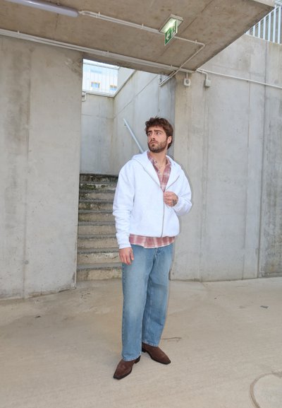 Hombre con sudadera blanca, camisa de cuadros, vaqueros y zapatos marrones de pie en una escalera de concreto bajo un cartel de salida, mirando hacia un lado.