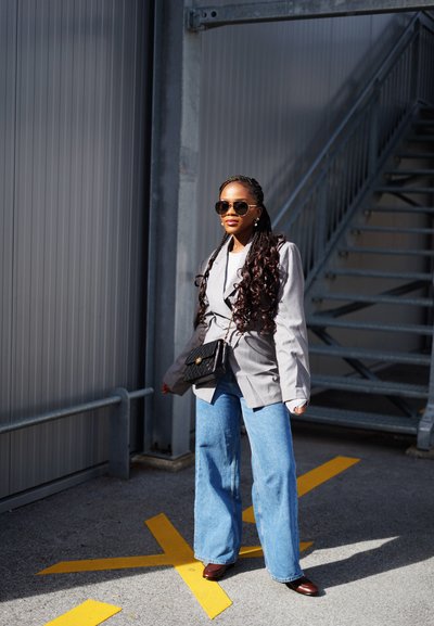 Blazer gris de gran tamaño sobre una camisa blanca, combinado con jeans de pierna ancha de color azul y zapatos marrones. Accesorios incluyen un bolso de hombro negro y gafas de sol.