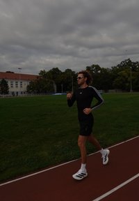 Camisa de treino de manga longa preta com riscas brancas, calções pretos e sapatos de corrida brancos com detalhes pretos, numa pista com relva por perto.