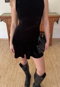 Robe en velours noir avec ourlet évasé, associée à des bottes noires montantes jusqu'aux genoux et un petit sac à main noir. Texture lisse, design simple.