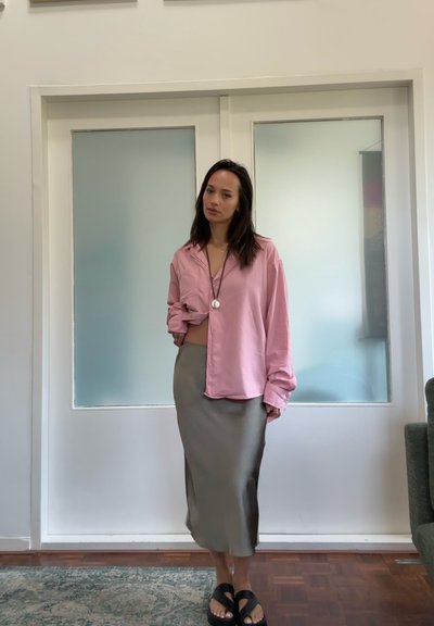 Camisa de satén rosa con botones y mangas enrolladas, combinada con una falda ajustada verde olivo y sandalias negras. Diseño minimalista y texturas suaves.