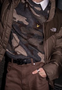 Veste matelassée marron portée sur un pull camouflage gris avec un petit emblème d'aigle doré, associée à un pantalon marron et une ceinture noire avec des trous circulaires.