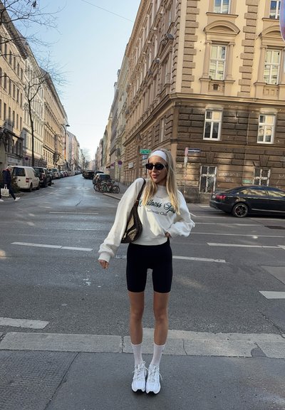 Sudadera blanca con texto verde, pantalones cortos de bicicleta negros, calcetines blancos y zapatillas chunky blancas. Bolso negro y gafas de sol. Fondo urbano.